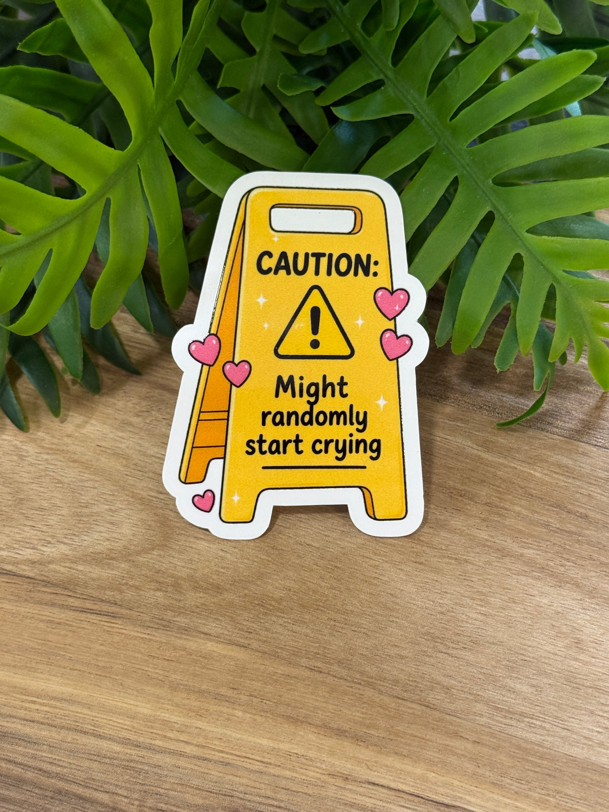 Yellow caution sign sticker with pink hearts reading Caution: Might randomly start crying