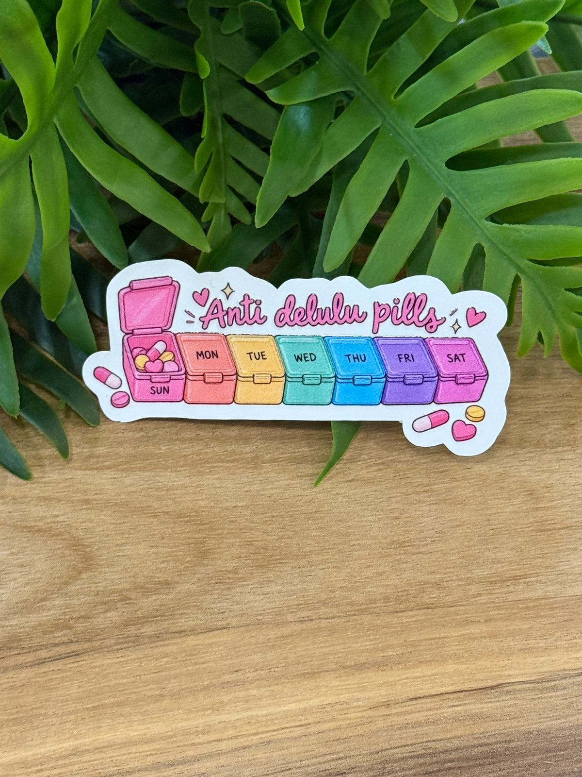 Anti delulu pills vinyl sticker featuring a pastel rainbow weekly pill organizer, pink hearts, illustrated pills, and sparkles