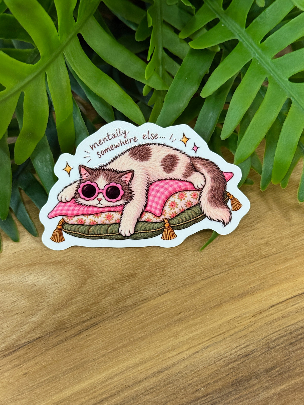 Mentally Somewhere Else cat sticker showing a fluffy cat wearing pink flower sunglasses and lounging on patterned cushions