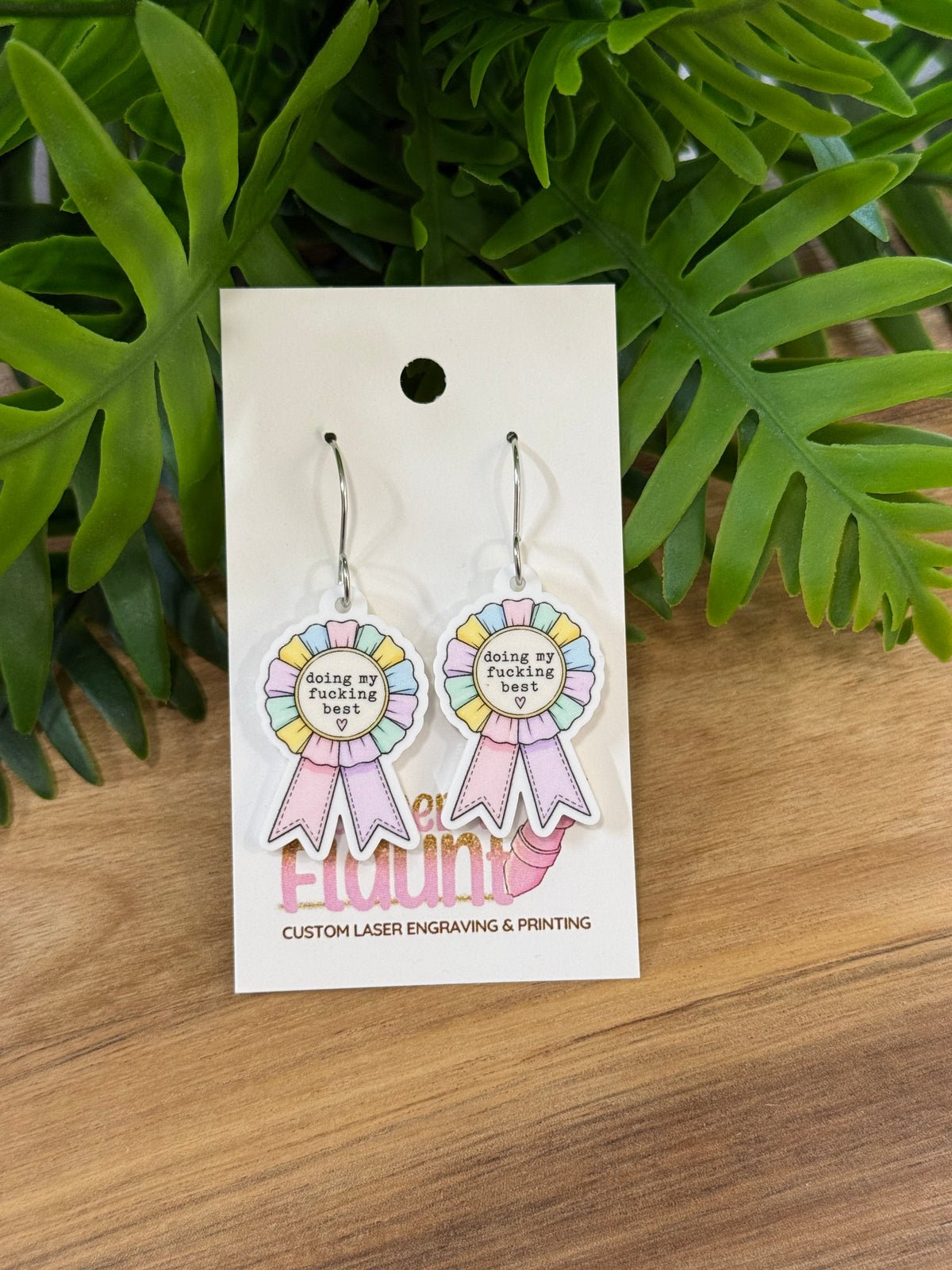 Pastel award-ribbon acrylic earrings reading doing my fucking best with pink and lavender ribbon tails and silver-colored wires
