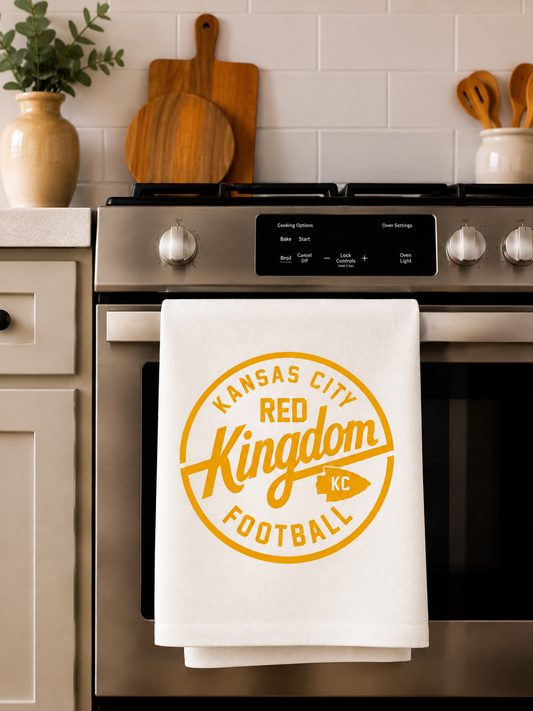 A white kitchen towel hanging on a stainless steel oven handle in a kitchen setting, printed with a yellow circular logo that reads 'Kansas City Red Kingdom Football'.
